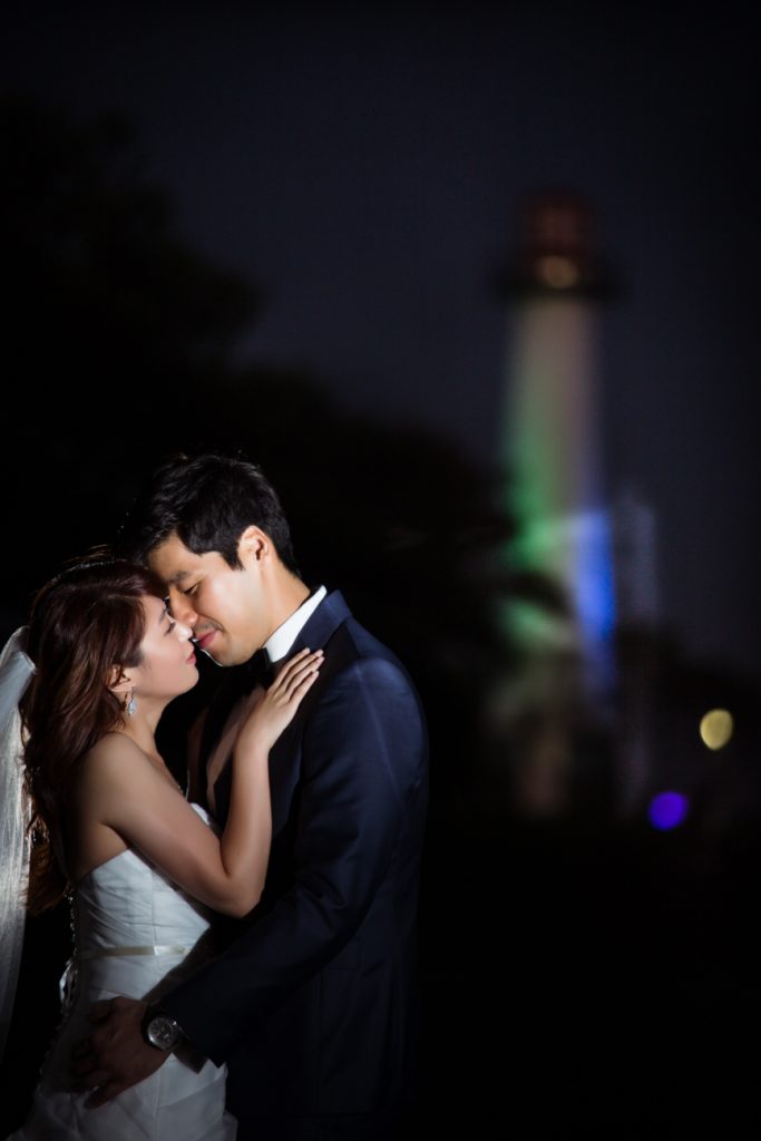 ThomasKim_photography A bride and groom embracing in front of a lighthouse at night, symbolizing their unity and strength.