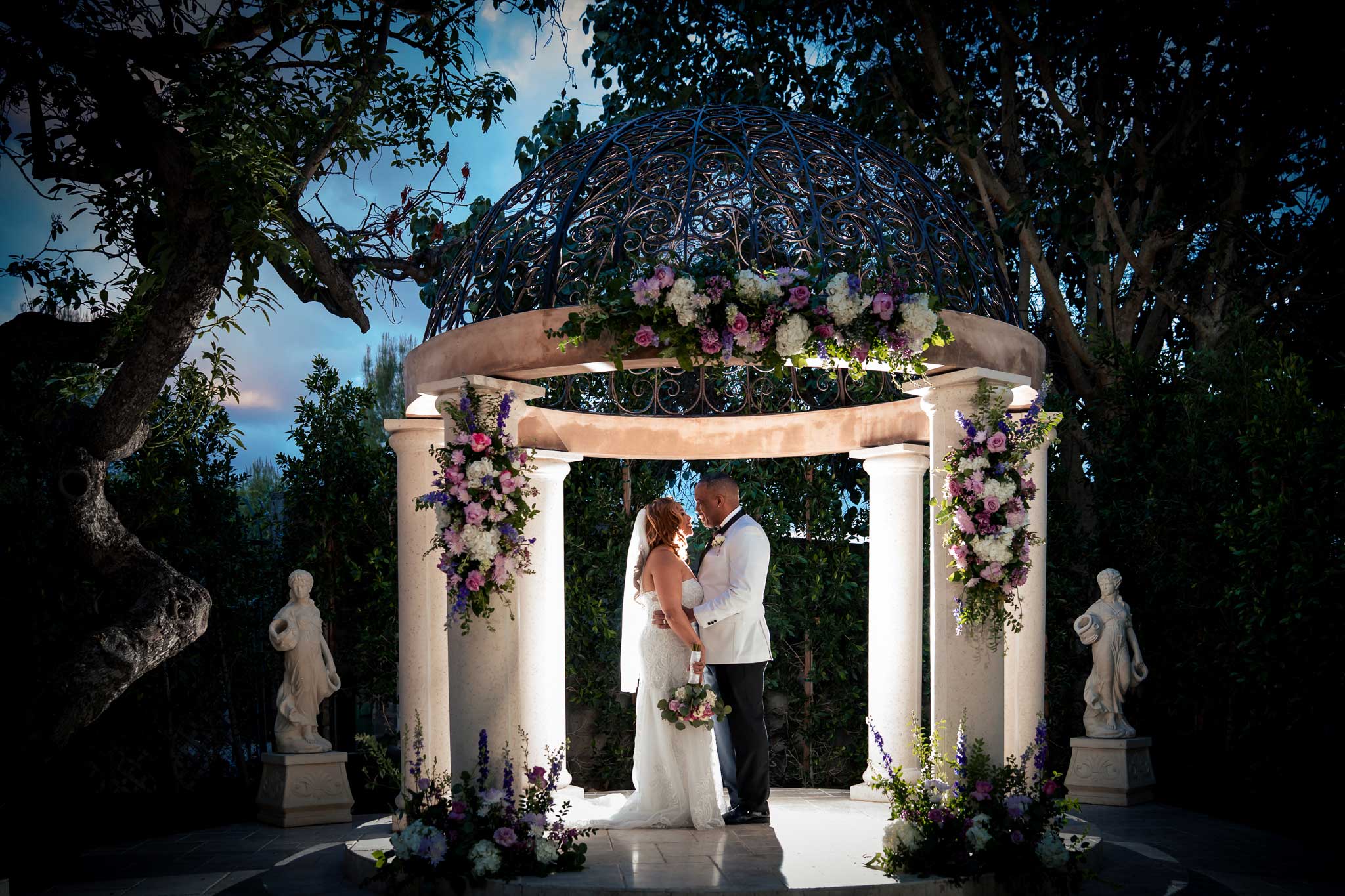 Bride and groom stand facing each other under a flower-decorated gazebo, surrounded by greenery and statues, during an outdoor wedding ceremony.