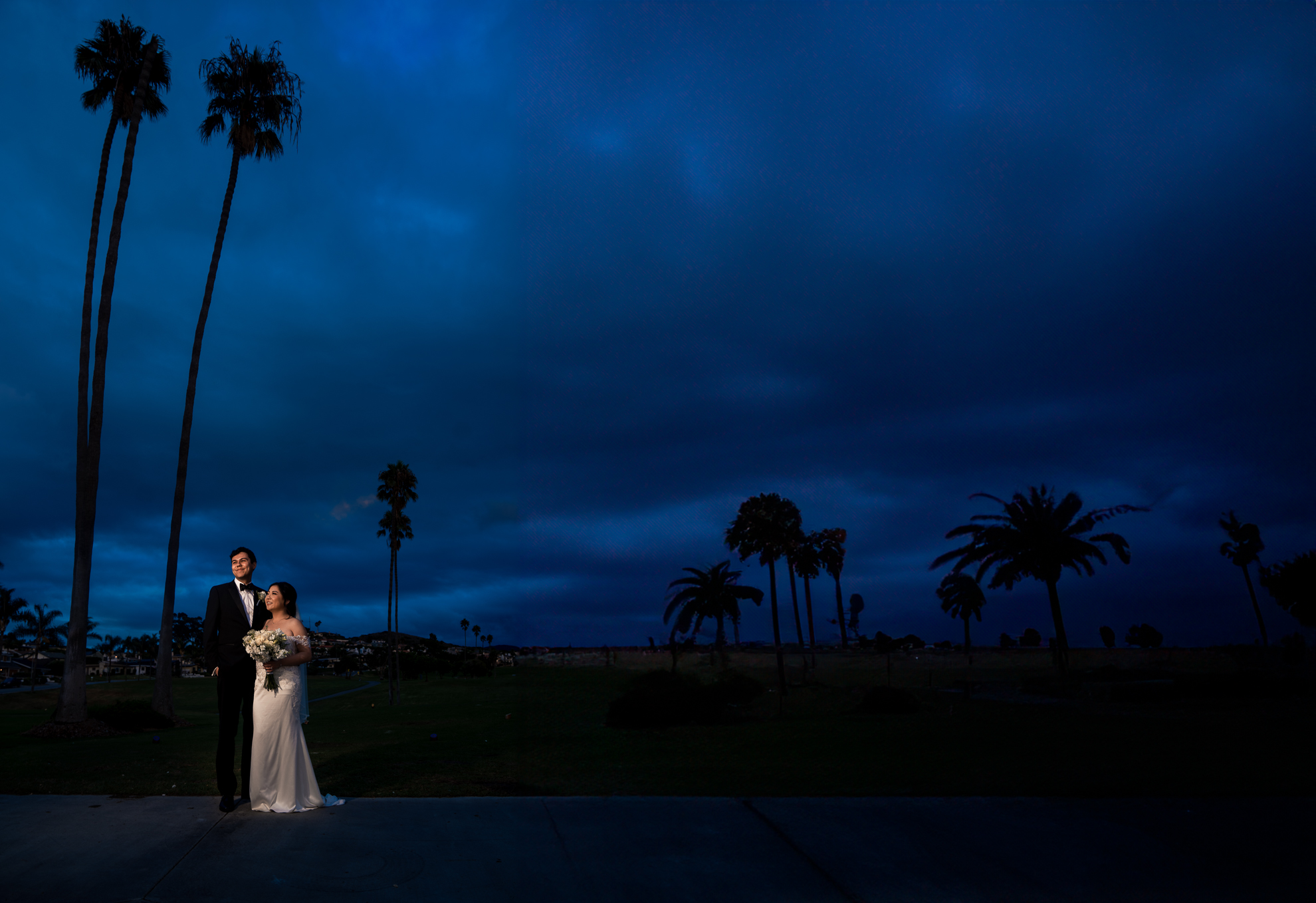 A bride and groom standing in front of palm trees at dusk.
