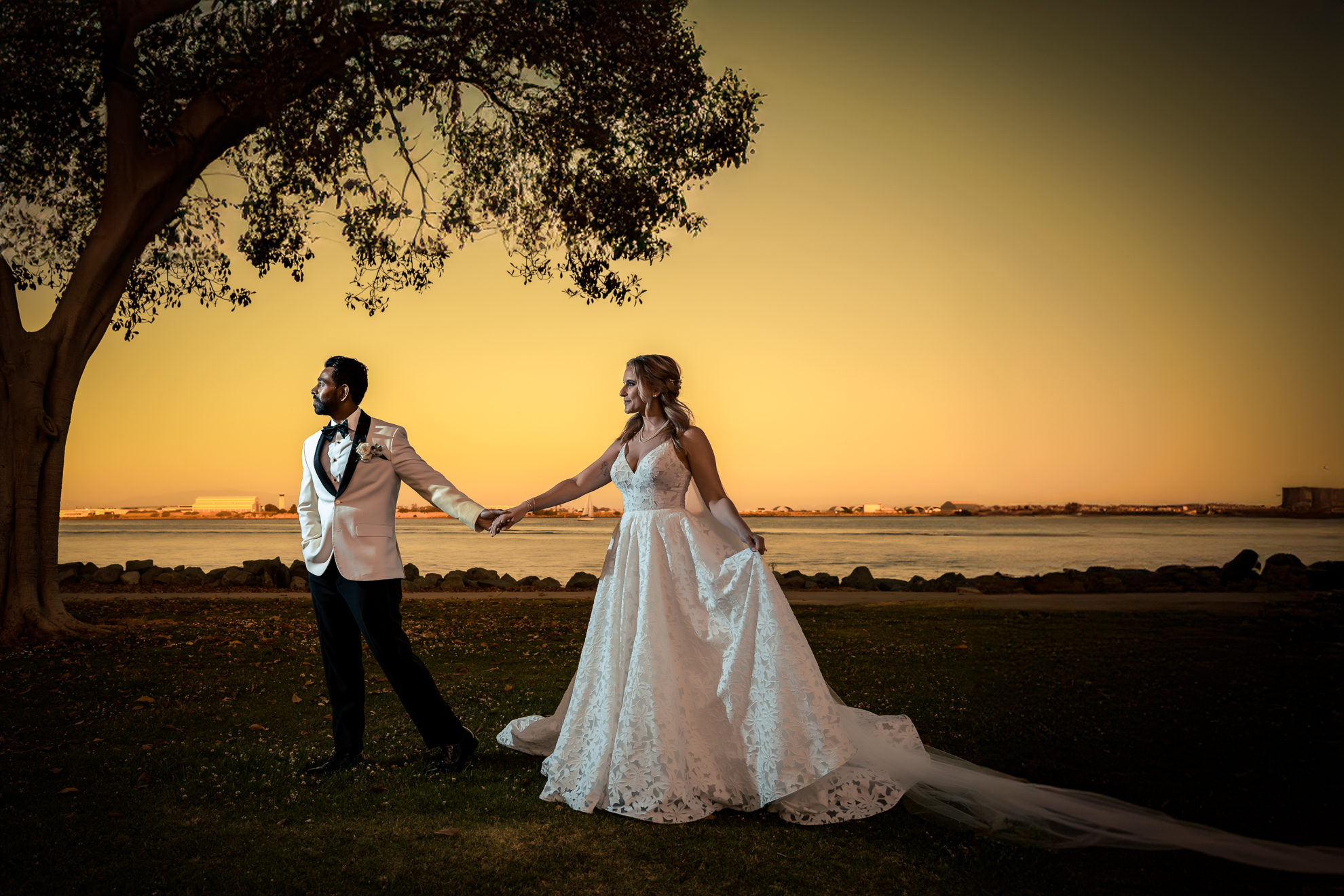 ThomasKim Photography Los Angeles Wedding Photographer A couple dressed in formal wedding attire holds hands under a tree at sunset by a waterfront. The bride wears a white gown with a long train, and the groom is in a white jacket and black pants.