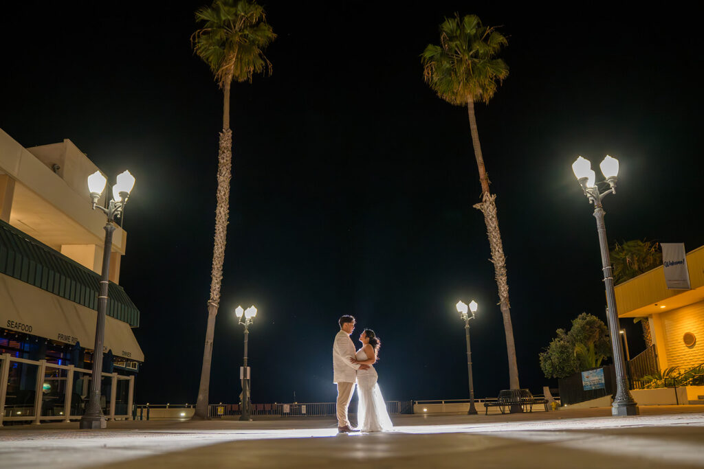 ThomasKim Photography Los Angeles Wedding Photographer A couple dressed in white stands close together at night, illuminated by streetlights, with two tall palm trees and buildings in the background.