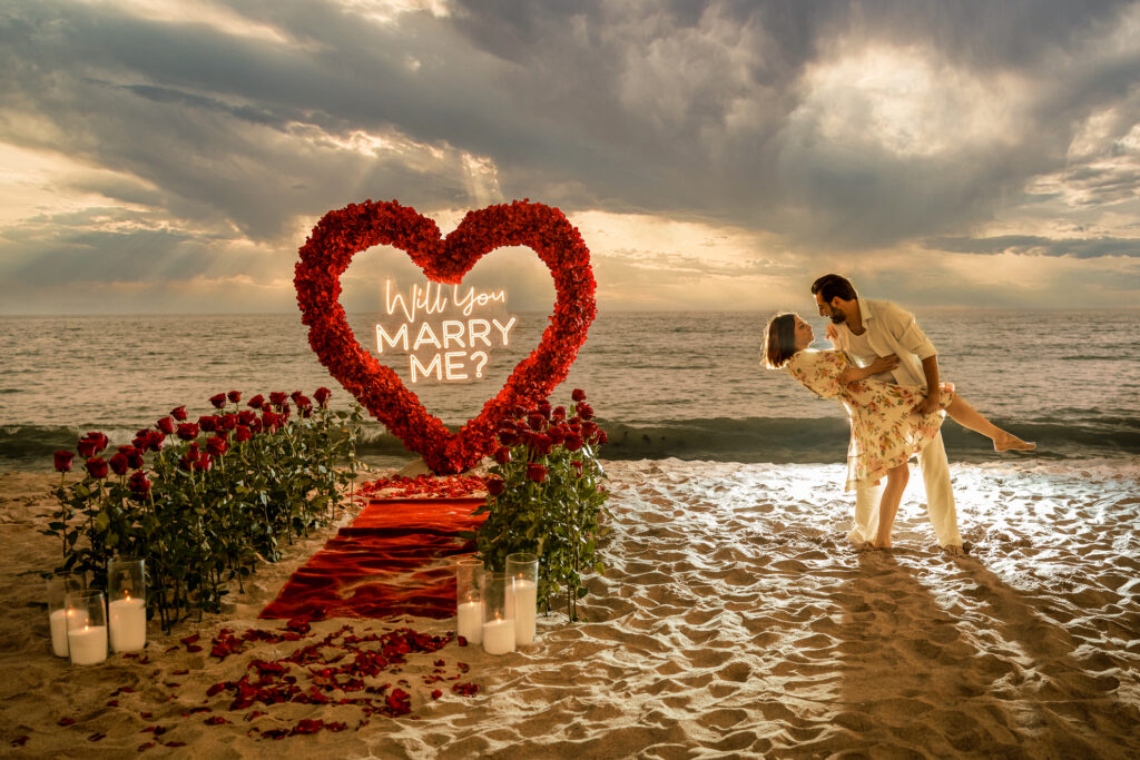 ThomasKim Photography Los Angeles Wedding Photographer A couple is posing in a romantic beach setting with a heart-shaped flower arrangement and candles. The flowers spell out "Will You Marry Me?" with a red carpet leading to the C & S Copy arrangement.