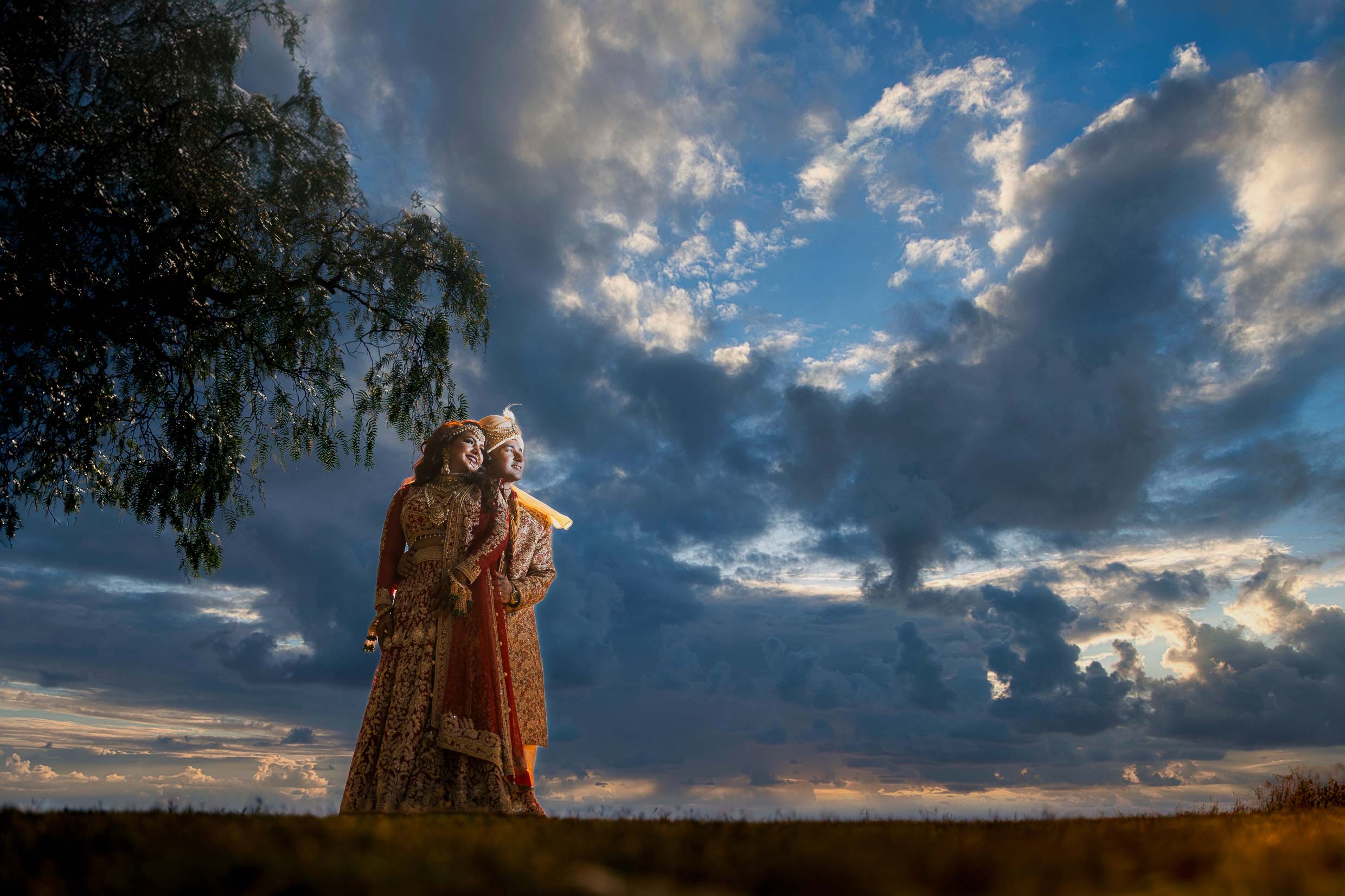 ThomasKim Photography Los Angeles Wedding Photographer A couple in traditional attire stands under a large tree against a dramatic, cloudy sky at sunset, captured perfectly by their wedding photographer in Los Angeles.