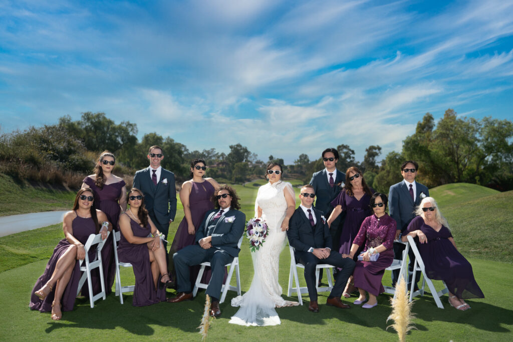 A bride and groom pose with ten guests in formal attire, seated and standing on a lawn under a blue sky, with trees and grass in the background.