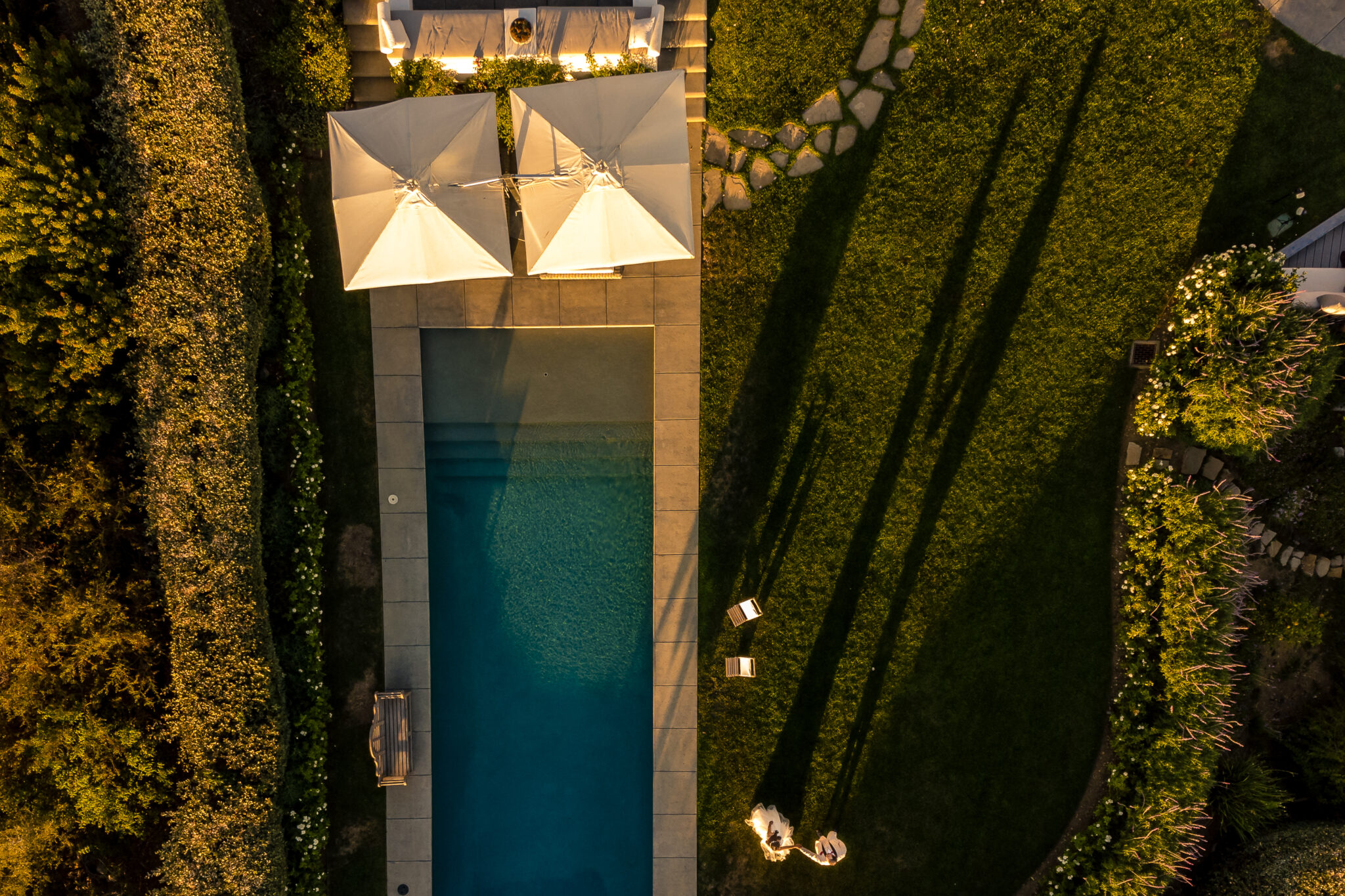 Aerial view of a rectangular backyard pool with two large umbrellas, lounge chairs, green lawn, and long shadows cast by two people.
