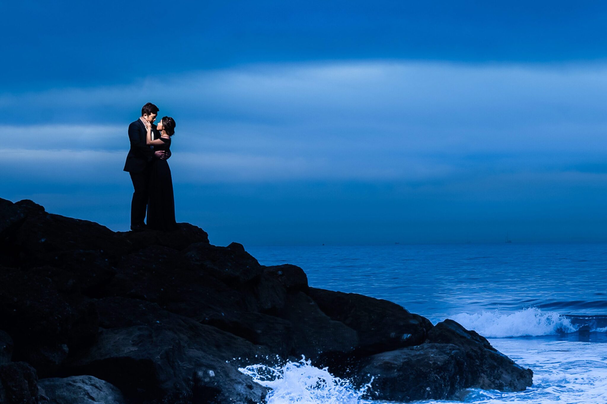 A couple in formal attire stands on rocky shore, embracing, with ocean waves crashing below and a blue sky overhead.