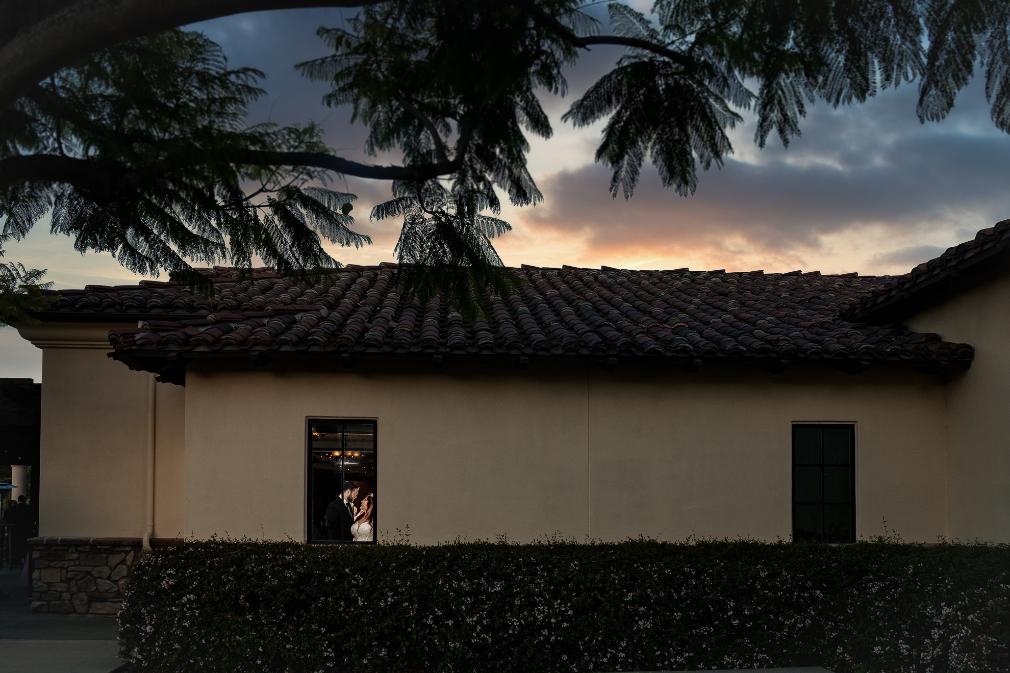 A beige building with a tile roof at sunset; people are visible through a lit window while trees frame the scene.