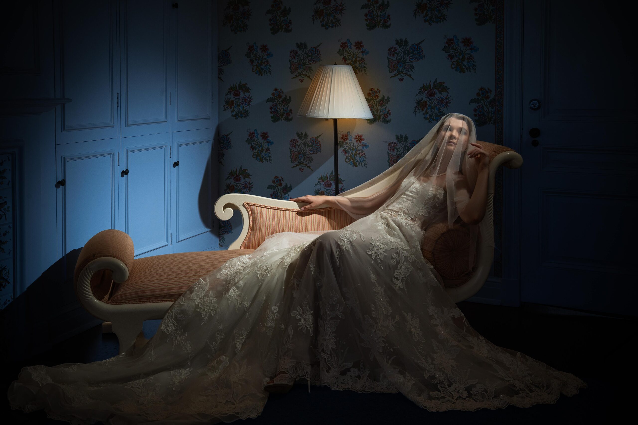 A bride in a long lace gown and veil reclines on a vintage chaise lounge in a dimly lit room with floral wallpaper and a lit floor lamp.