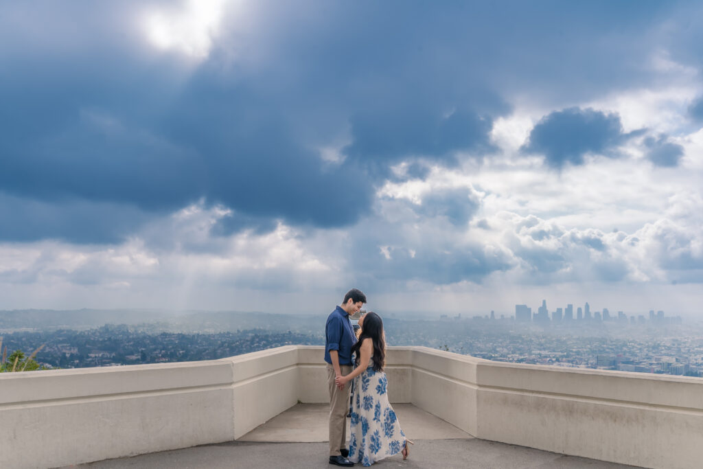 A couple stands close together on a scenic overlook with a city skyline and dramatic cloudy sky in the background.