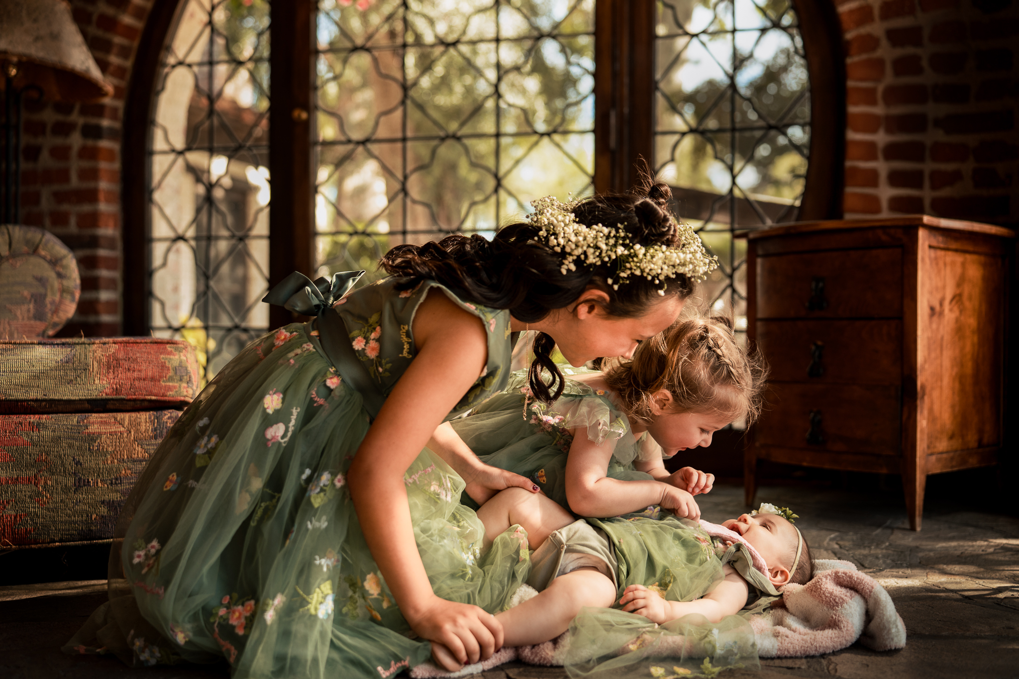 Three young girls in green floral dresses play together on the floor in a sunlit room with large windows and brick walls.