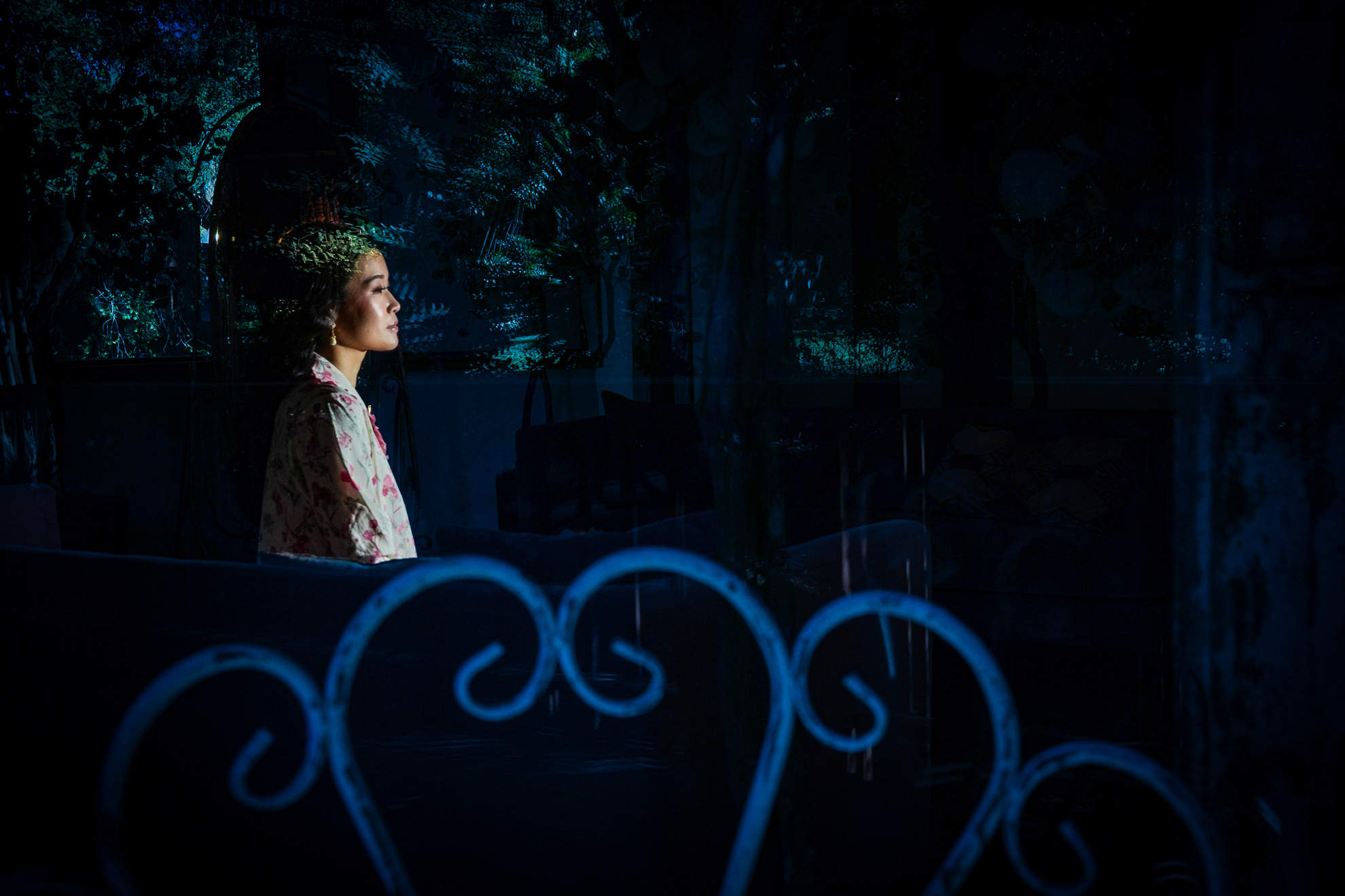 A woman in a floral shirt sits indoors, illuminated by blue-tinted light, with swirling patterns and reflections visible in the foreground.