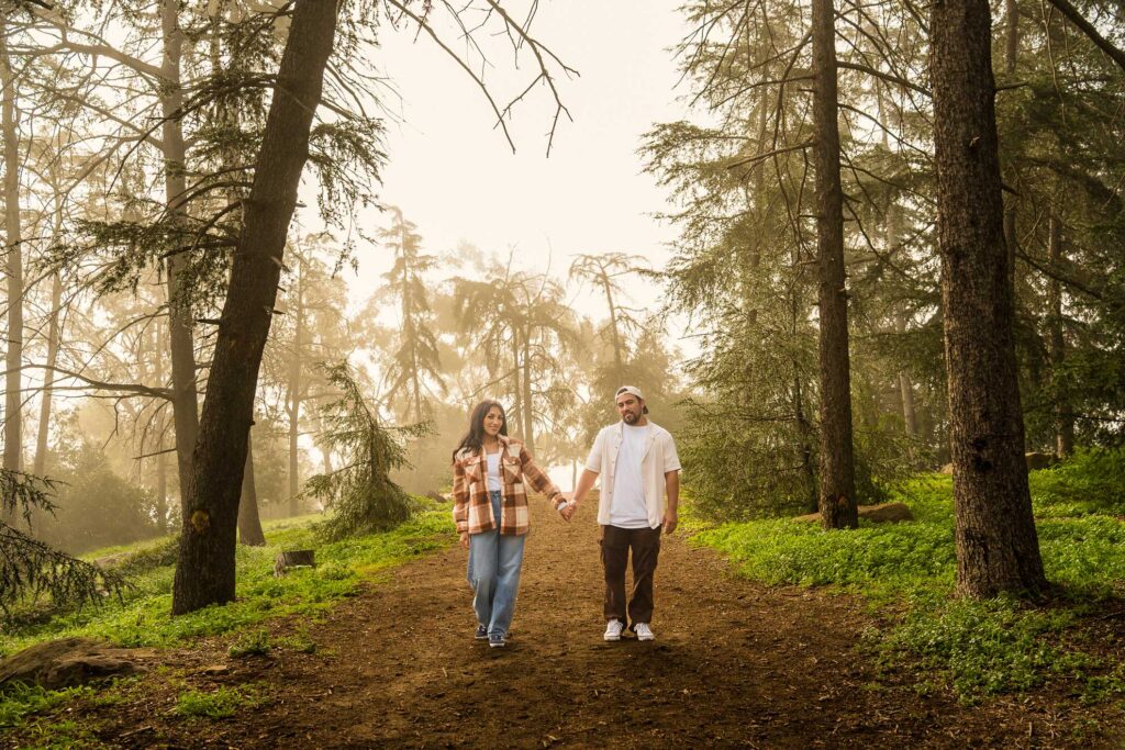 A man and woman hold hands while walking on a dirt path through a forest with sunlight filtering through the trees.