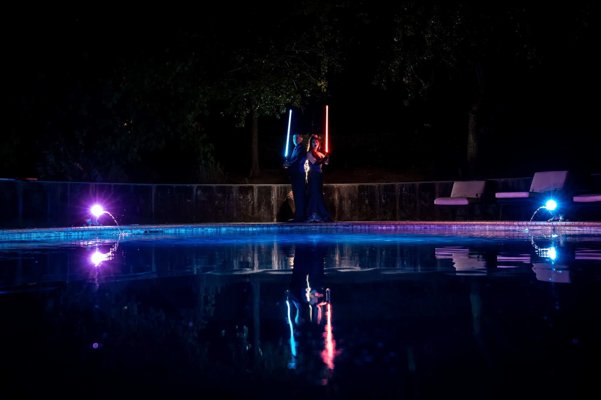 Two people stand by a pool at night holding illuminated lightsabers, one blue and one red, with reflections on the water and colorful lights around them.