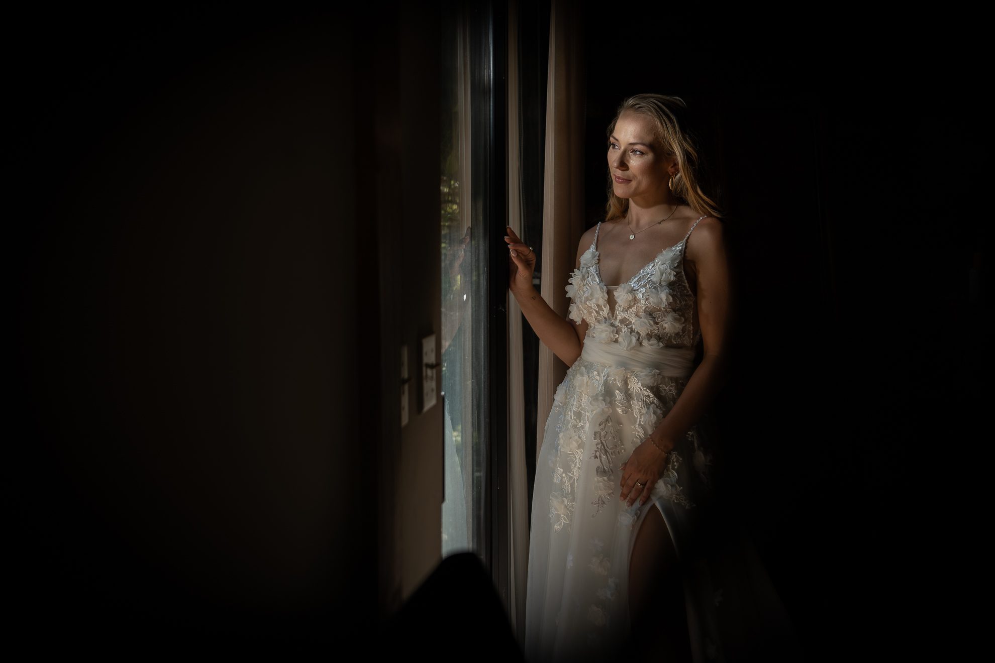 A woman in a white, floral wedding dress stands by a window, looking outside with a soft smile. Light from the window illuminates her face and dress, while the background remains dark.