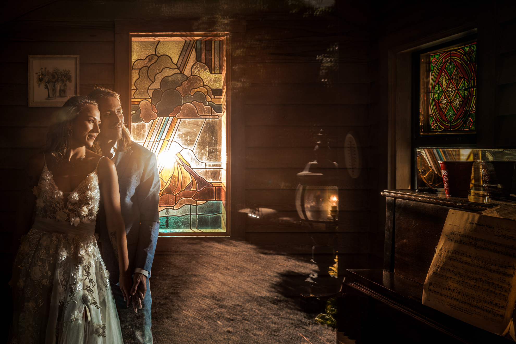 A bride and groom stand in a dimly lit room with stained glass windows, sunlight streaming in, and sheet music on a nearby piano.