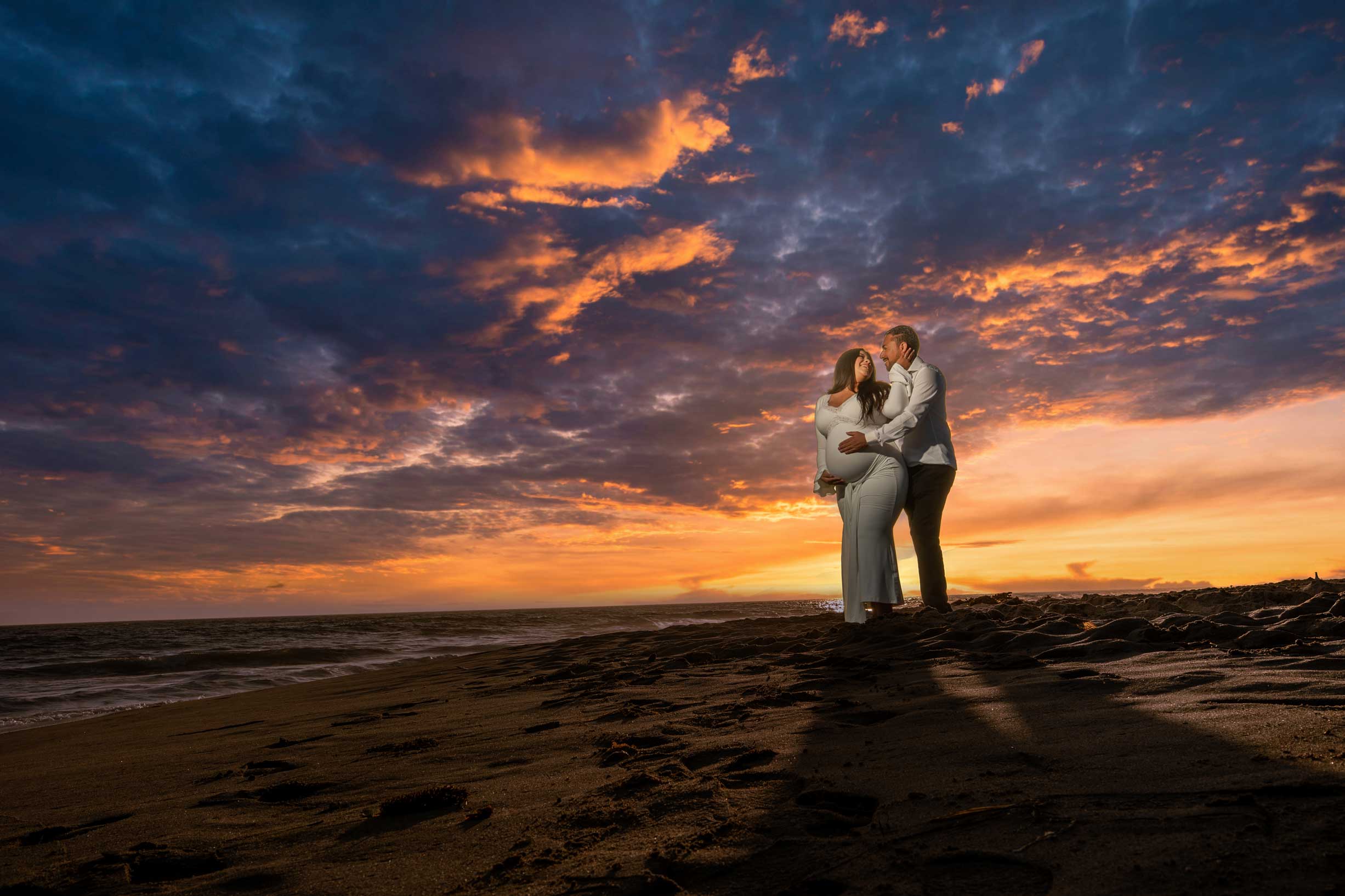 A couple embraces on a sandy beach at sunset, with dramatic clouds and colorful sky in the background.