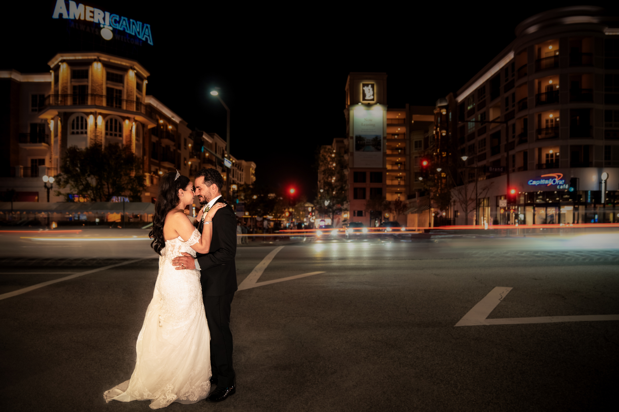 A bride and groom in wedding attire embrace on a city street at night, surrounded by illuminated buildings and blurred car lights.
