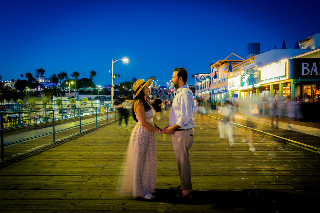 A couple stands facing each other and holding hands on a busy boardwalk at dusk, with blurred people passing by and lit buildings in the background.