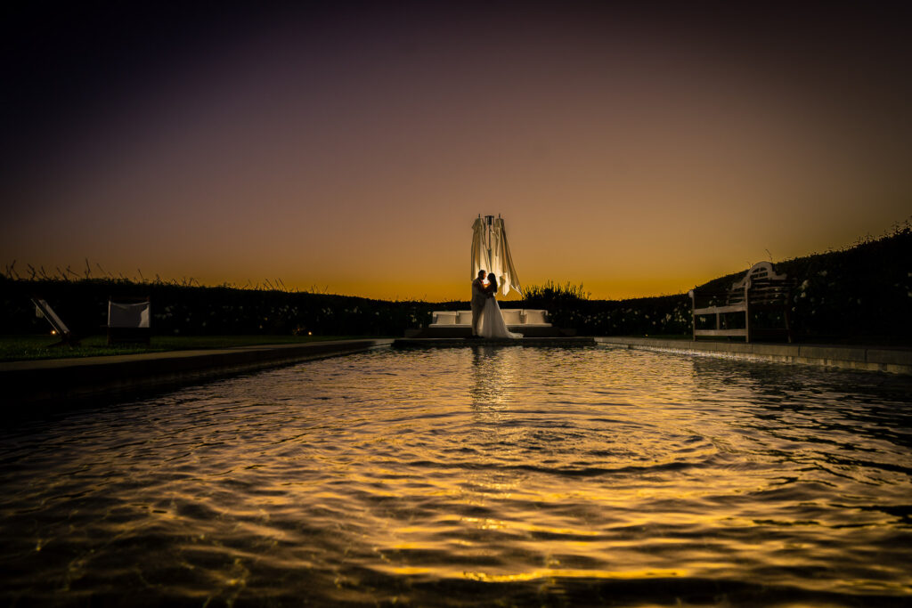 ThomasKim_photography A newlywed couple standing in front of a beautiful fountain during a mesmerizing sunset.|ThomasKim_photography A monochrome photo of two individuals seated in a room.|ThomasKim_photography A wedding dress hung from an exquisite olive tree.|ThomasKim_photography A pair of silver shoes with crystals on them