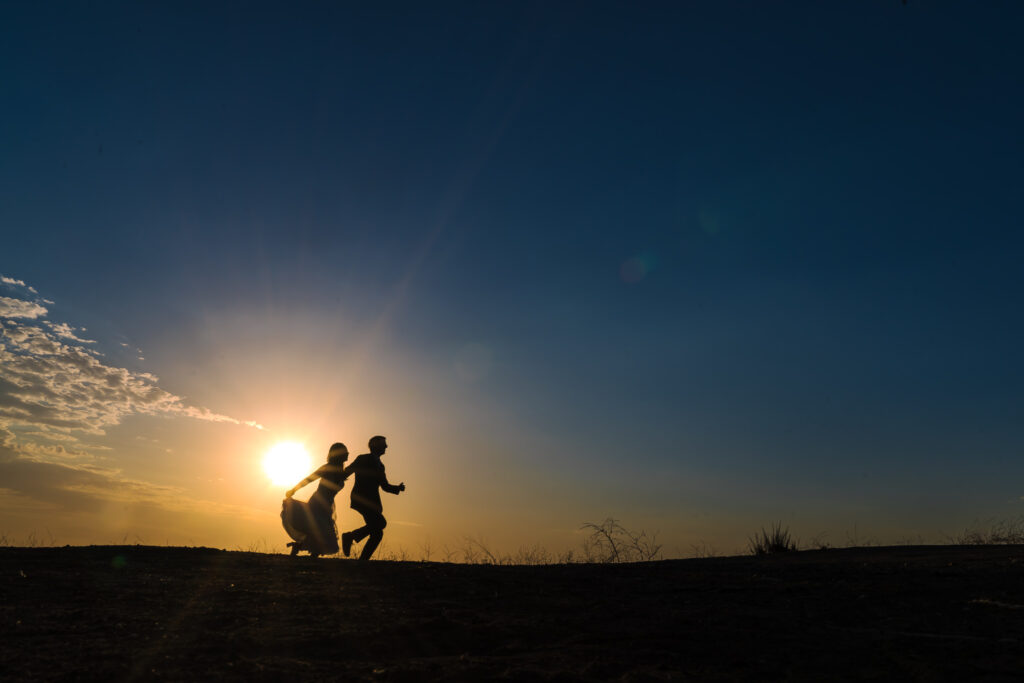 Silhouettes of two children, one crouching and one running, against a sunset sky with scattered clouds and visible sun near the horizon.