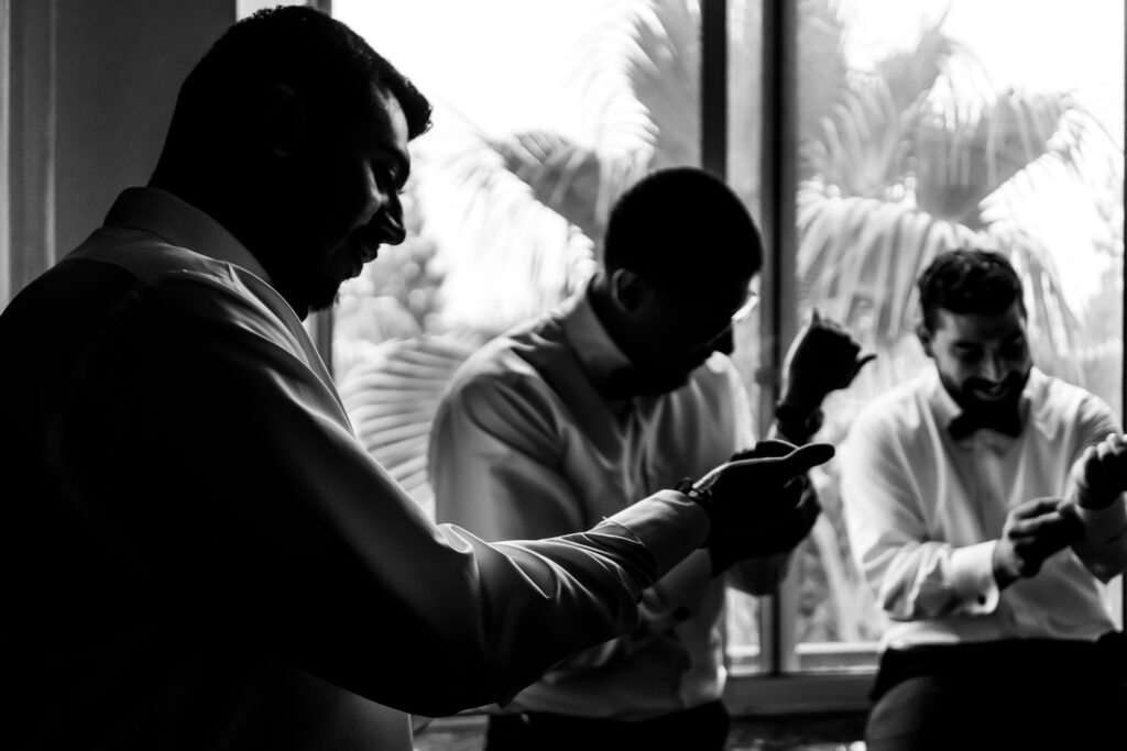 Three men in dress shirts adjust their cuffs and sleeves near a window with palm trees visible outside, shown in black and white.