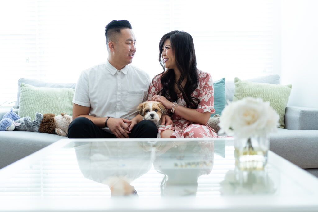 A couple sits on a sofa with a small dog between them, smiling at each other. Plush toys and pillows are on the couch, and a rose in a vase is on the table in the foreground.