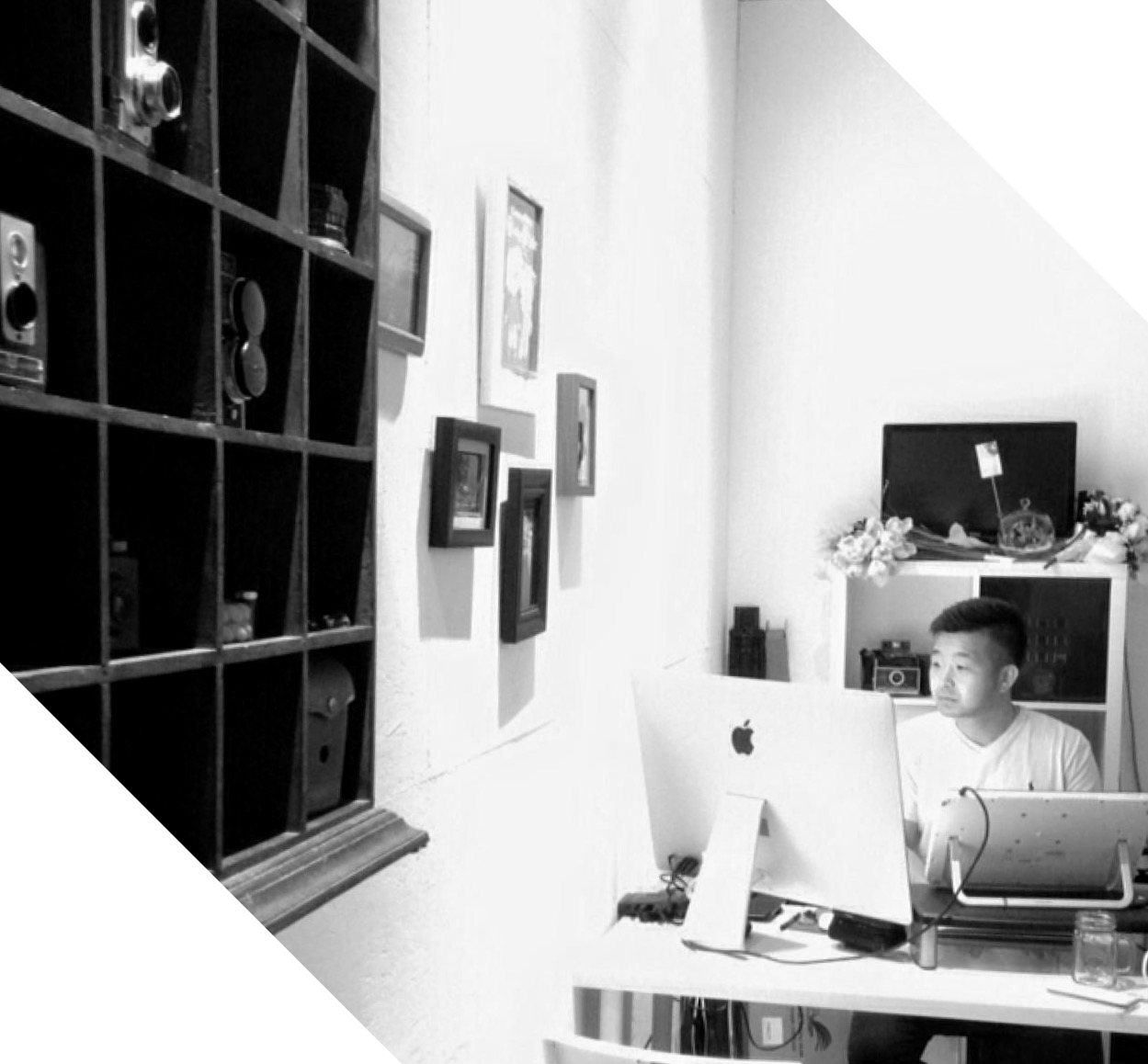 A man sits at a desk with an imac in a room decorated with framed pictures and a wall shelf holding vintage cameras.