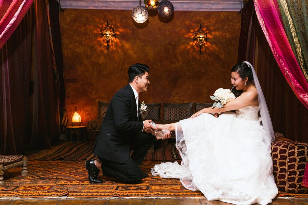 A wedding photographer captures a Los Angeles bride and groom as they put on their shoes in a room.|A bride posing for a photographer in front of a rack of wedding dresses.|Thomas Kim