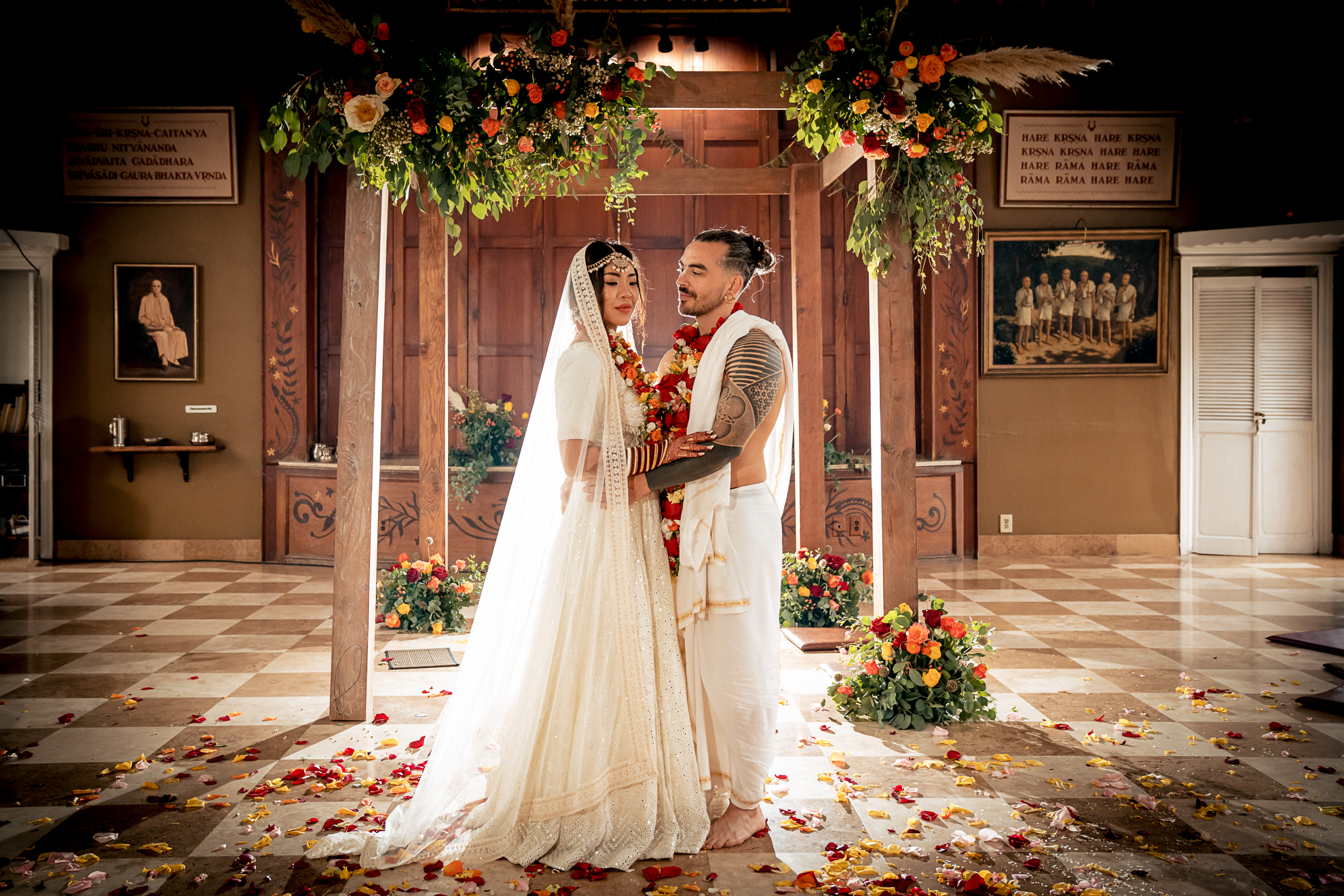 A luxury bride and groom stand in traditional attire beneath a floral canopy in a beautifully decorated indoor venue, surrounded by scattered flower petals, creating timeless portraits of elegance.