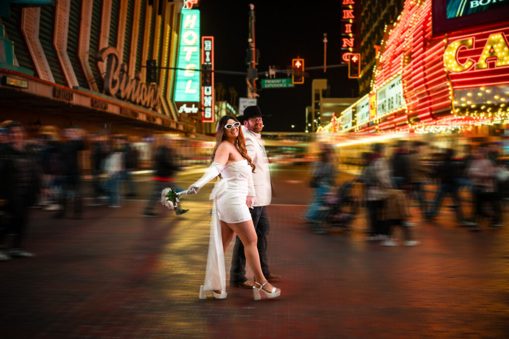 A couple in wedding attire poses on a lively city street at night, surrounded by blurred crowds and neon lights.