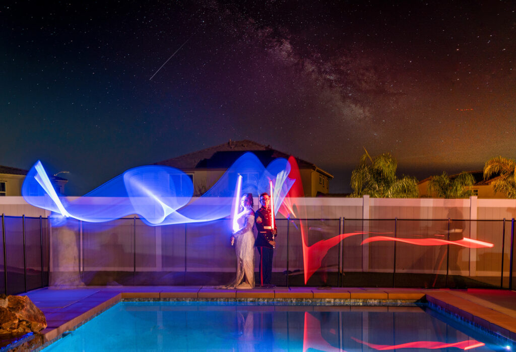 A couple in formal attire stands by a pool at night, holding lightsabers with red and blue light trails, under a starry sky with visible houses and palm trees in the background.