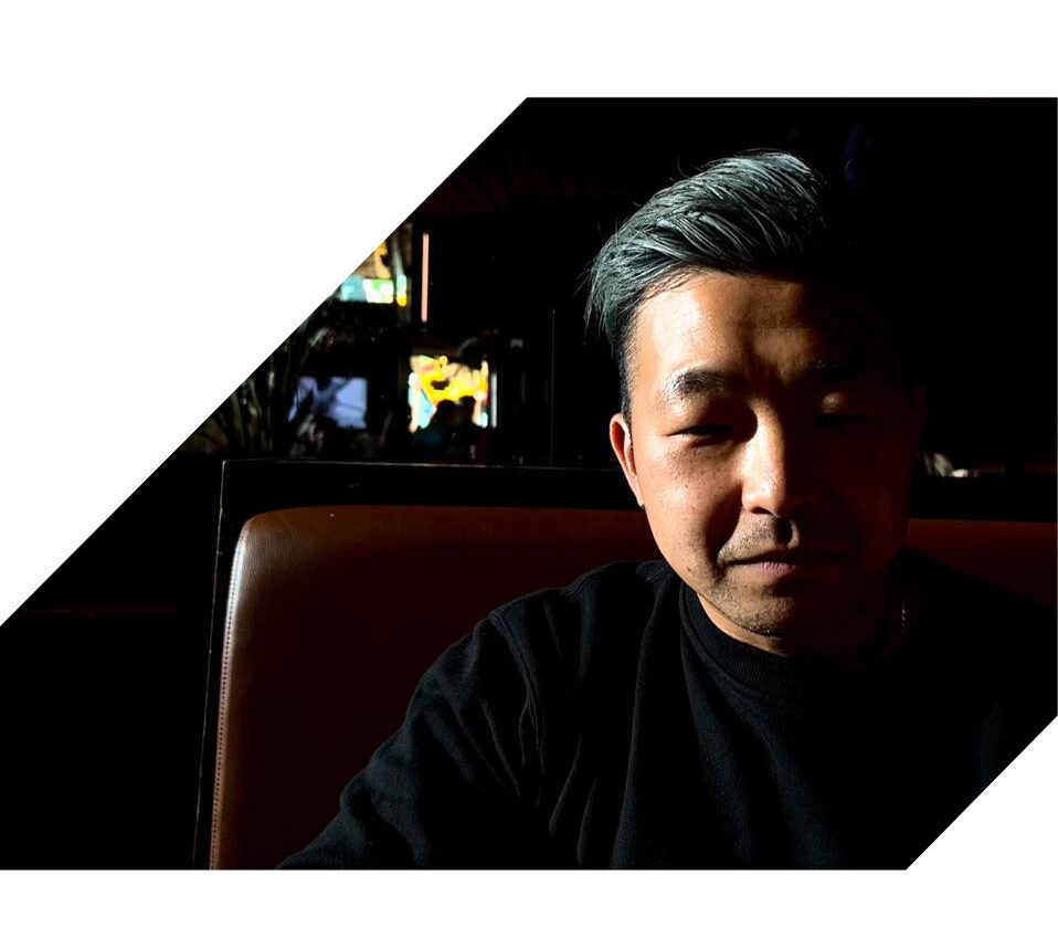 ThomasKim Photography Los Angeles Wedding Photographer A man with gray hair sits in a dimly lit booth, with half of his face illuminated. The background is dark, and a bright white geometric shape frames the left side of the image.