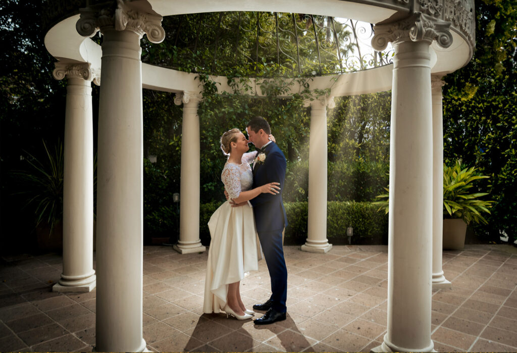 ThomasKim Photography Los Angeles Wedding Photographer A couple in wedding attire embraces under a decorative gazebo surrounded by greenery