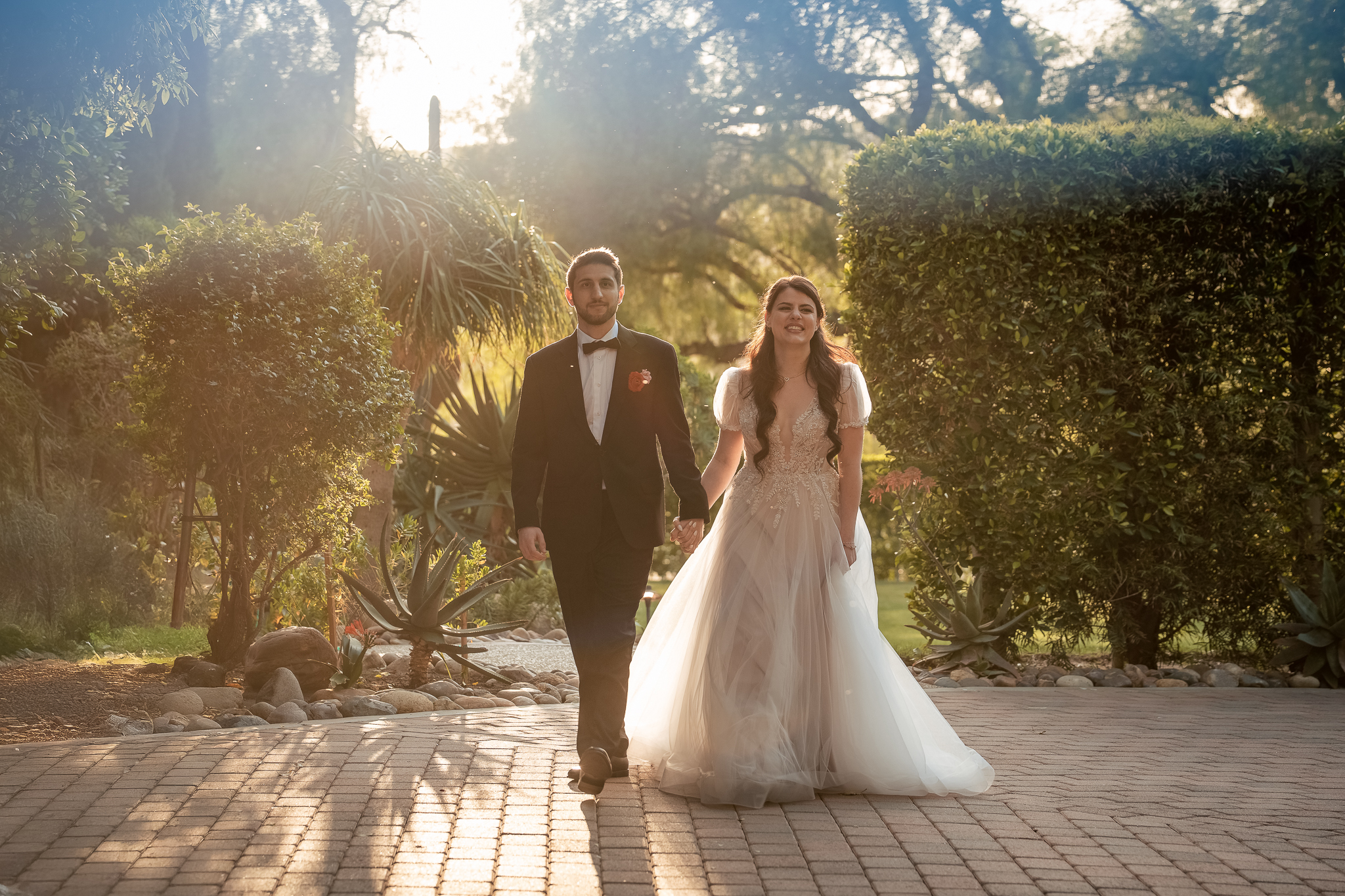 A bride and groom walk hand in hand outdoors on a sunny day, surrounded by greenery and soft sunlight.