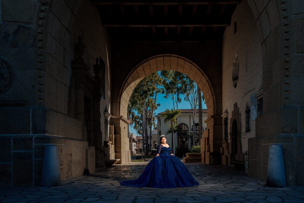 A woman in a blue ball gown stands under a large stone archway, with sunlight and trees visible in the background.