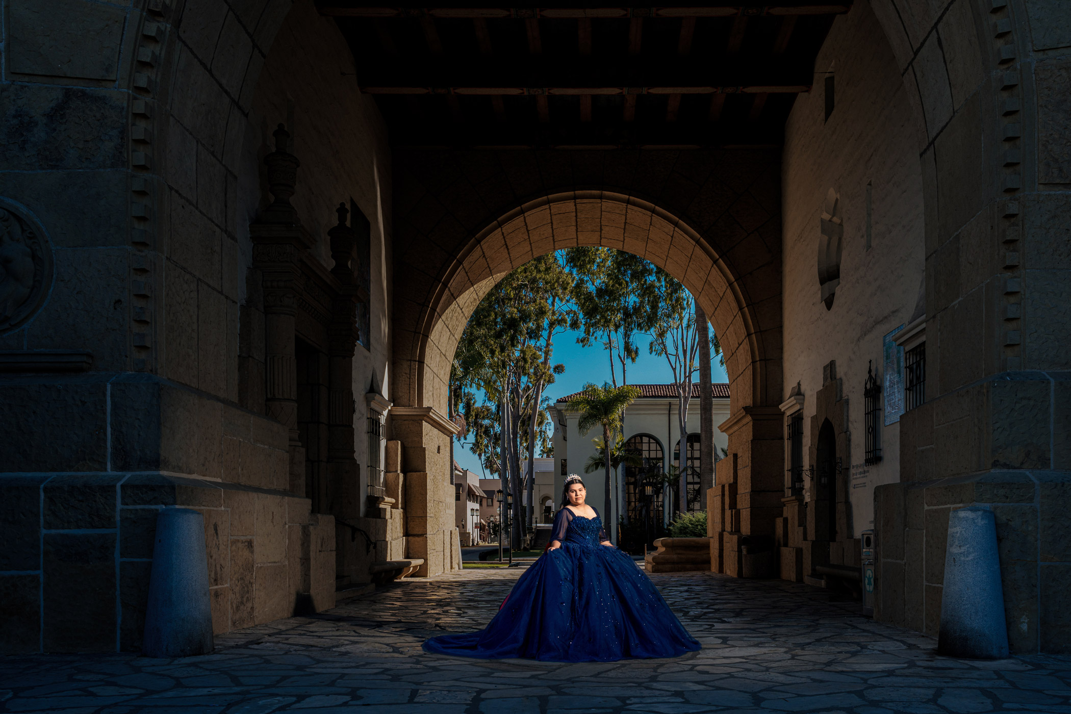 A woman in a blue ball gown stands under a large stone archway, with sunlight and trees visible in the background.