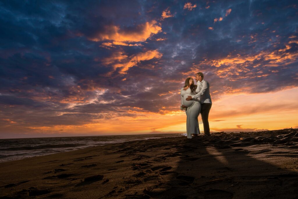 A couple stands embracing on a sandy beach at sunset, with dramatic clouds and vibrant orange and purple colors in the sky.