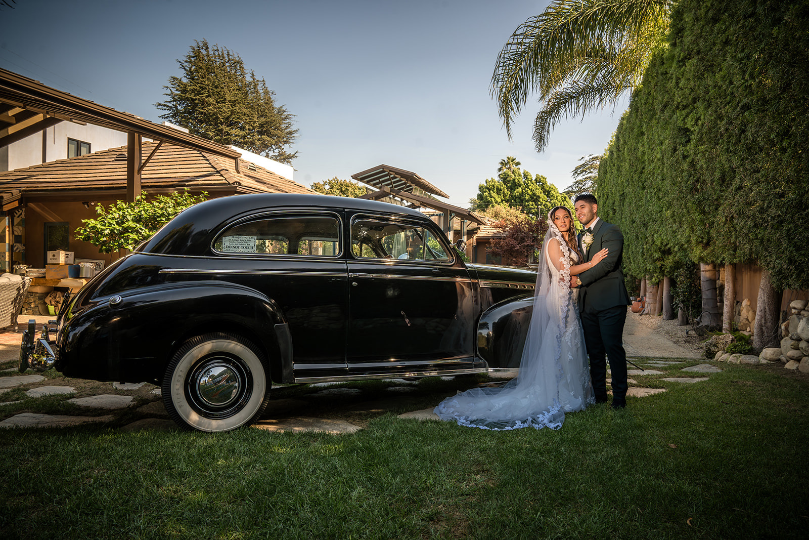 A bride and groom stand beside a vintage black car in a grassy backyard, surrounded by trees and a house under a clear sky.