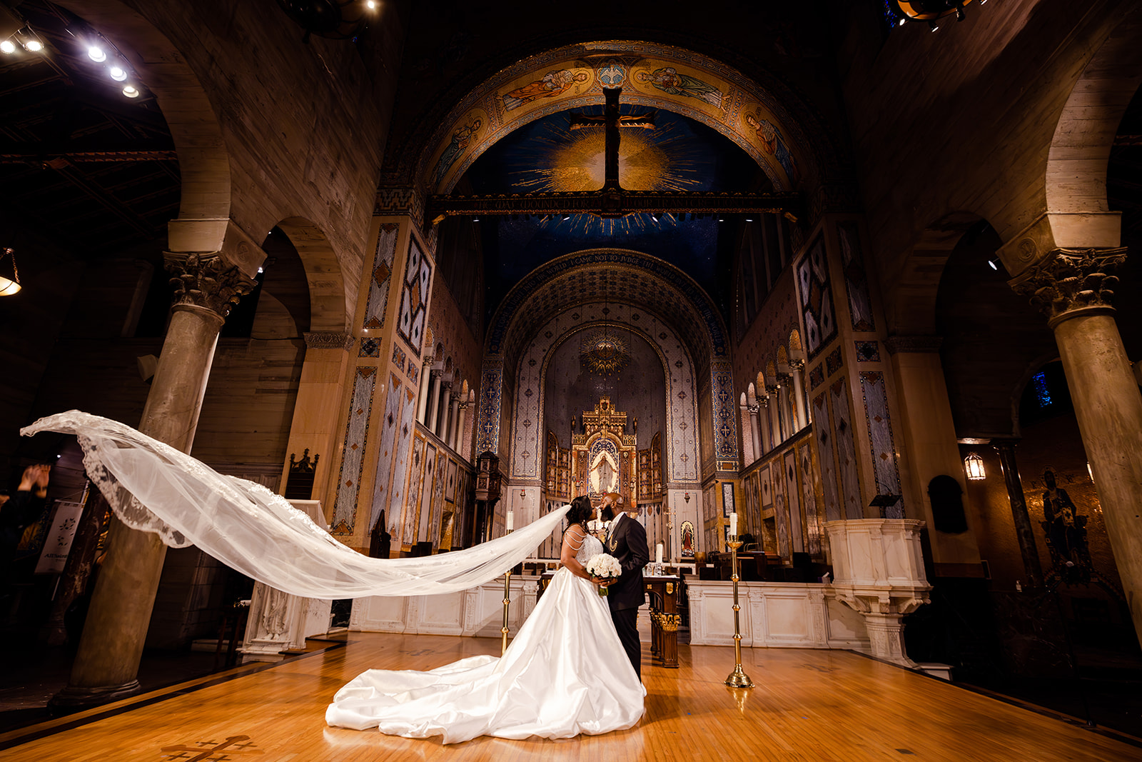 A bride and groom kiss at the altar of an ornate church; the bride’s long veil flows behind her on the wooden floor.