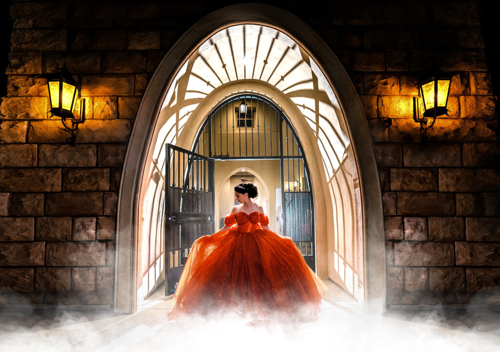 A person in an orange gown stands in an arched stone doorway, with dramatic lighting, open gates behind them, and lamps on the wall. Mist covers the ground in the foreground.