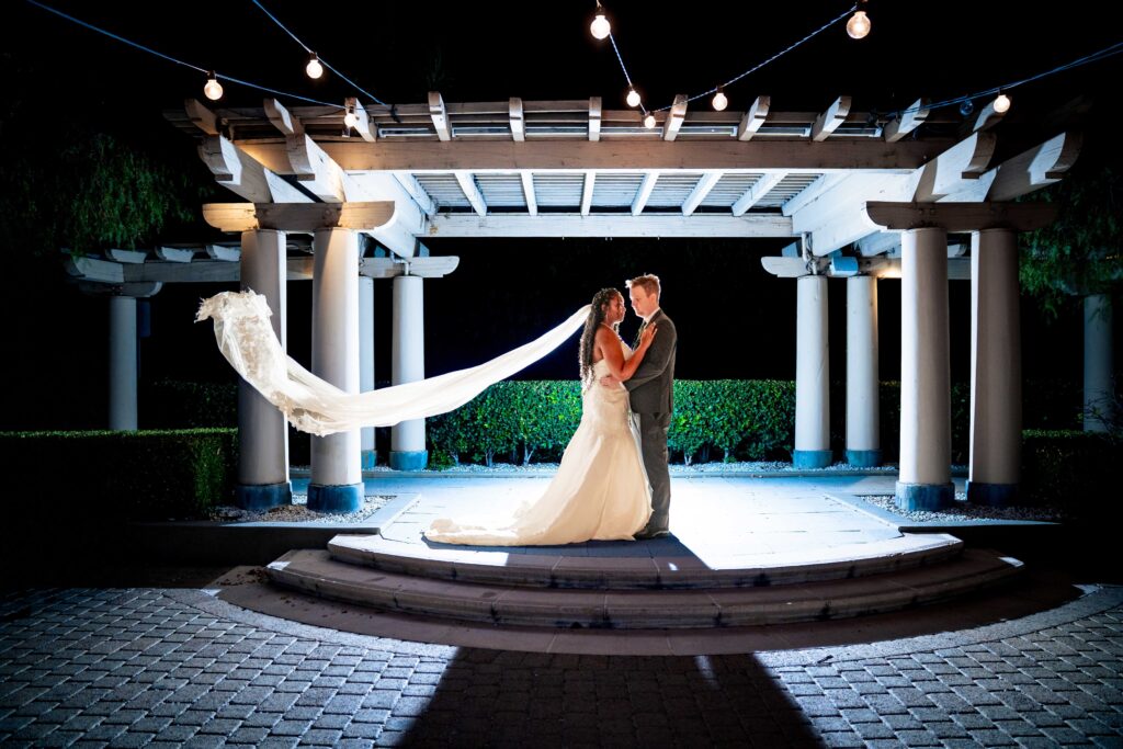 A bride and groom stand together under a lit pergola at night, with the bride’s veil flowing behind her and string lights overhead.