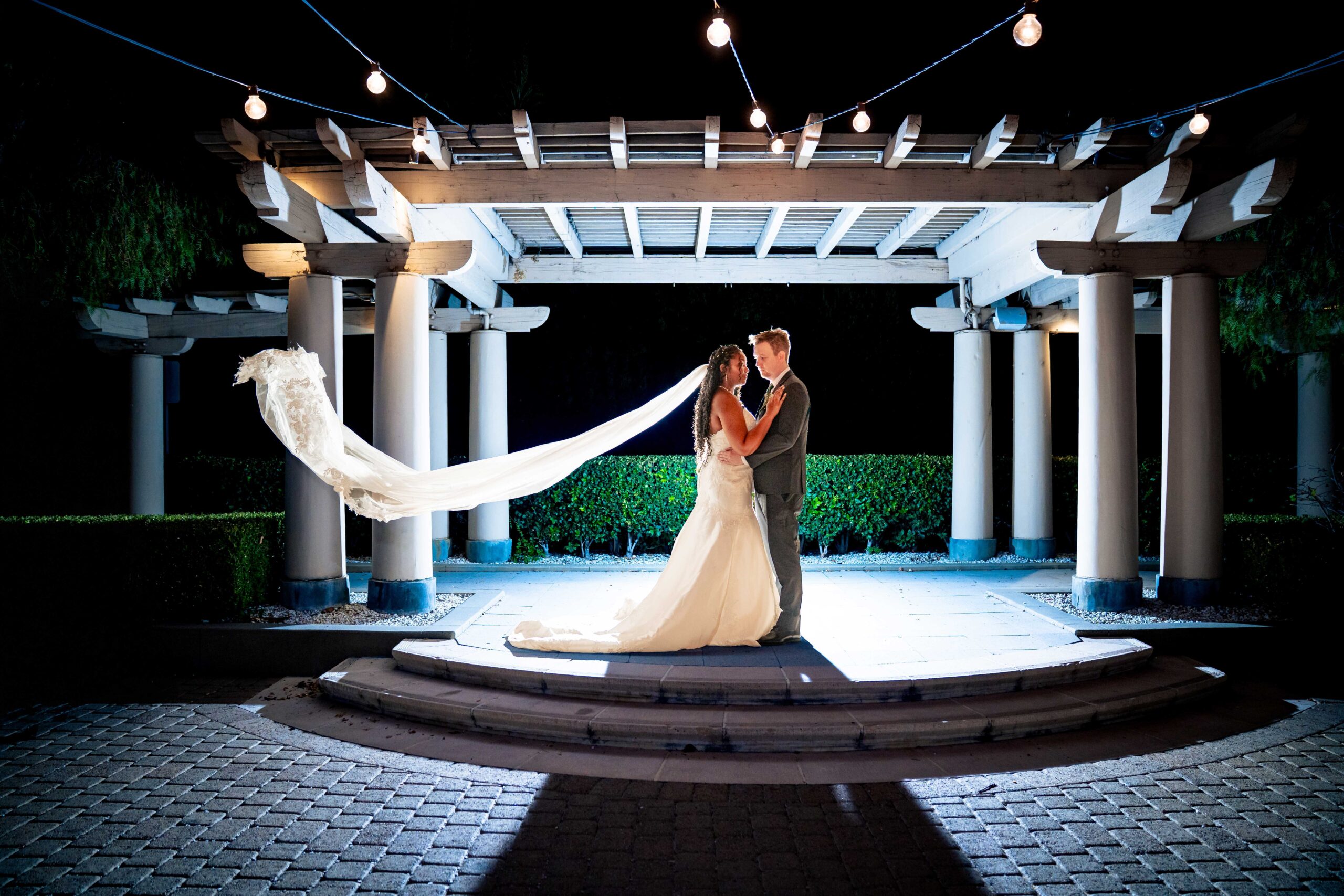 A bride and groom stand together under a lit pergola at night, with the bride’s veil flowing behind her and string lights overhead.