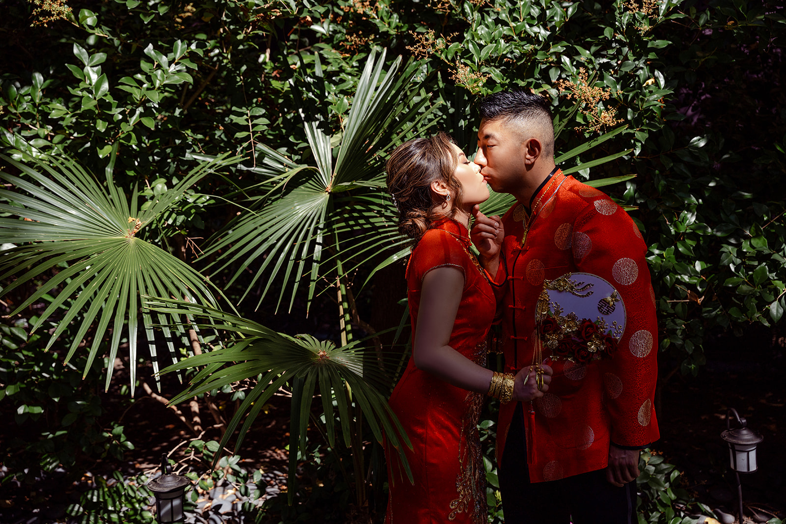 A couple in traditional red attire shares a kiss outdoors, surrounded by green foliage and palm leaves.