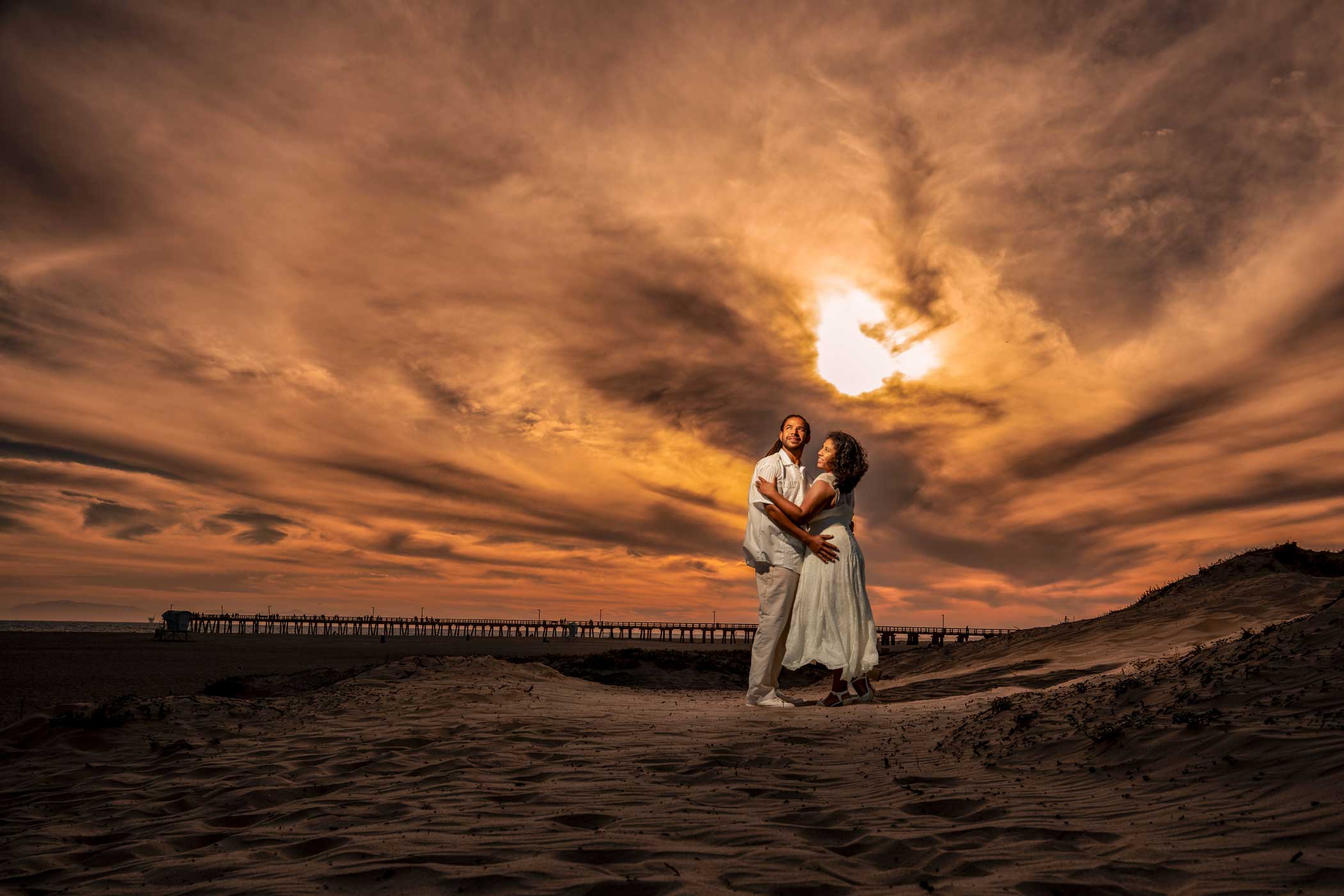 A couple in white clothing stands on sandy ground at sunset, embracing under a dramatic, cloud-filled sky with a pier in the background.