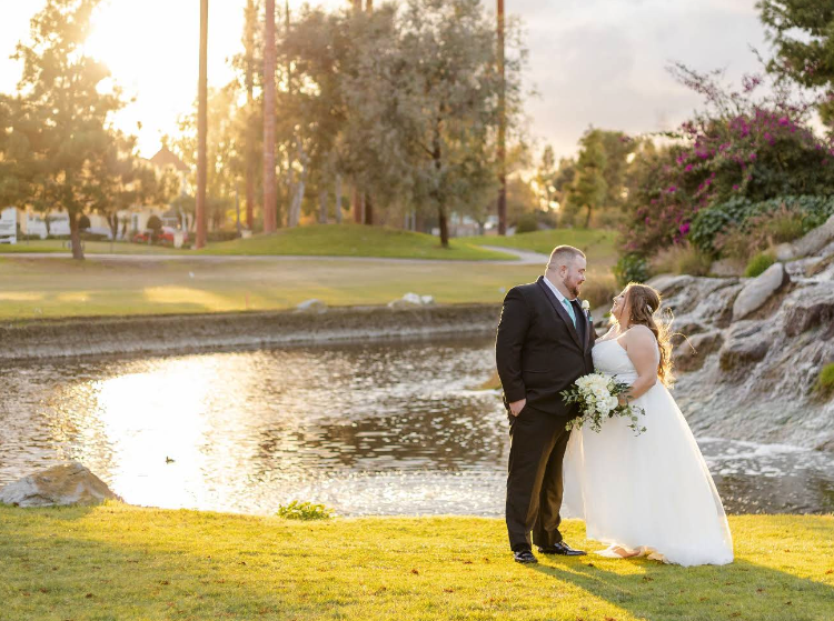 A bride and groom stand together by a pond at sunset, holding a bouquet, with trees and grass in the background.