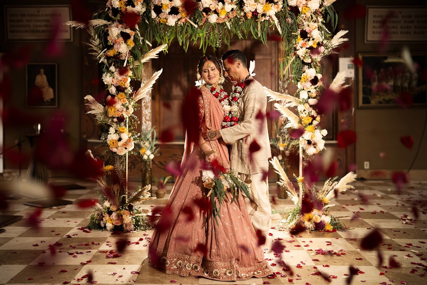 A luxury bride and groom stand together under a flower arch indoors, wearing traditional wedding attire, with rose petals falling around them—creating an elegant setting perfect for timeless luxury bride and groom portraits.