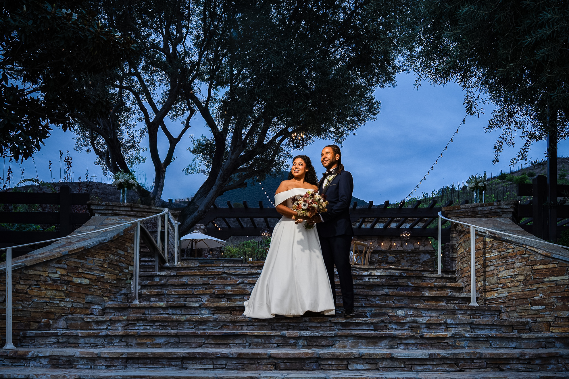 A bride and groom stand together on a stone staircase at dusk.
