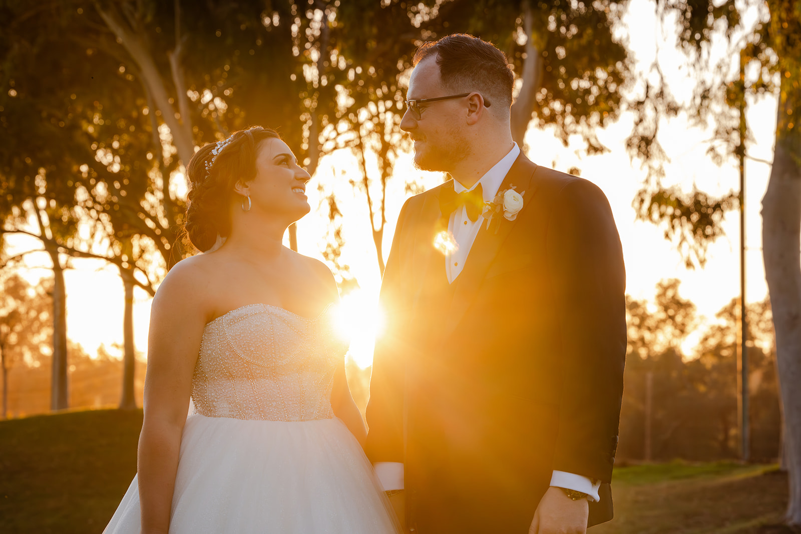 A bride and groom standing in a park at sunset, looking at each other.